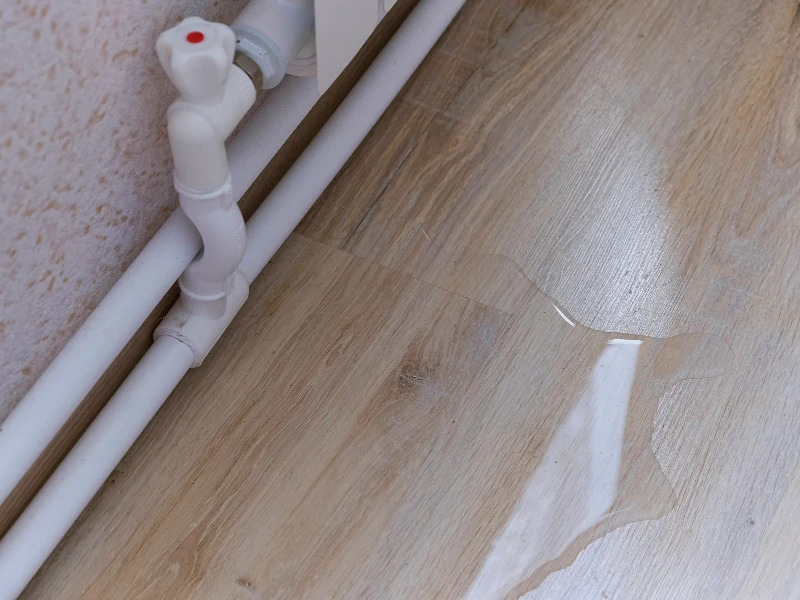 Water leak pooling on a wooden floor near white plumbing pipes and a valve. leak detection services